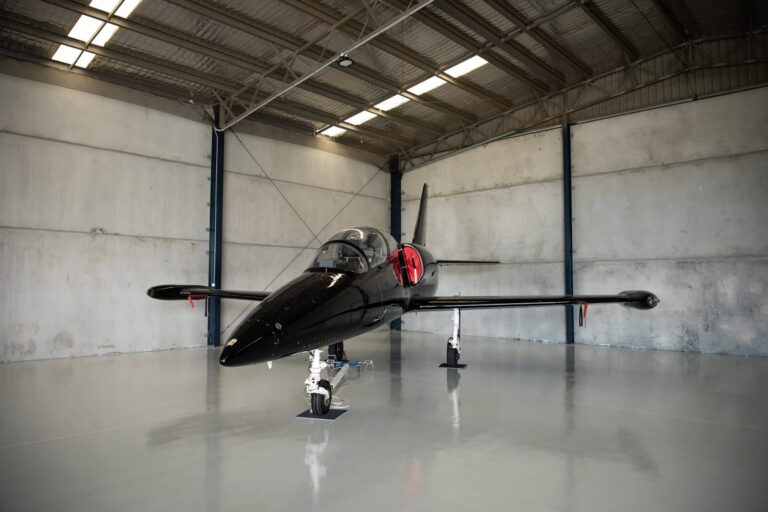 Black jet trainer nose-on inside an empty hangar bay with concrete panel walls