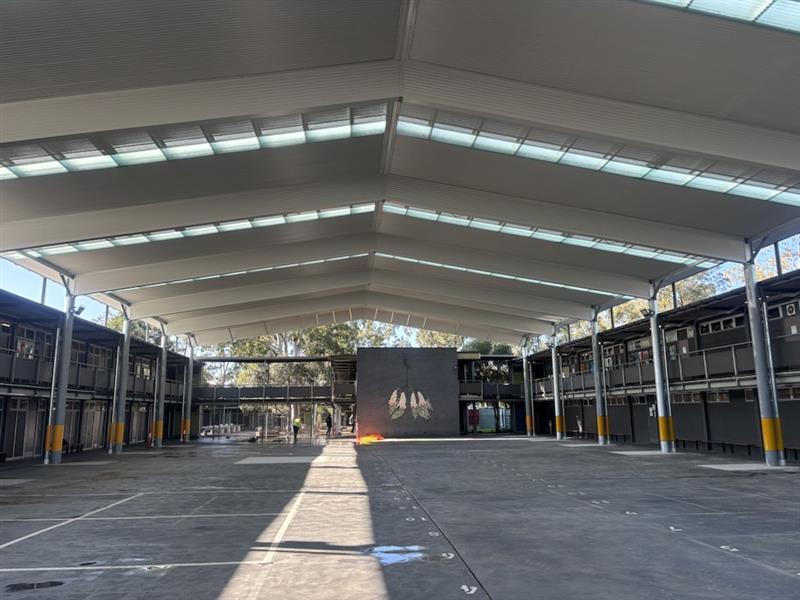 Large clear-span steel COLA structure covering a school quadrangle with translucent roof panels allowing natural light into the covered outdoor space.